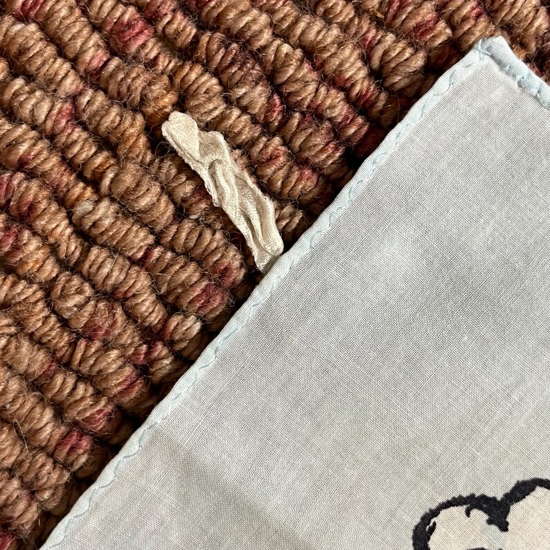 May include: A light blue handkerchief with a light blue border and a decorative design. The handkerchief is partially visible, resting on a textured brown and red surface. A small, cream-colored fabric piece is also present.