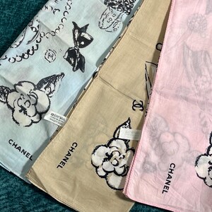 May include: Three folded square scarves in light blue, tan, and pink. Each scarf features a floral design, the word "CHANEL", and other graphic elements. The scarves are made of a soft fabric.