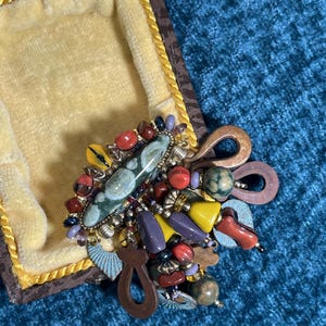 May include: A vintage brooch with a cluster of colorful beads and stones. The brooch features a large, oval green stone at the center, surrounded by red, yellow, purple, and blue beads. The brooch is set in a gold-tone metal frame.