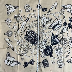 May include: Beige scarf with a repeating pattern of black and white illustrations. The design includes high heels, camellias, bows, and pearl necklaces. The word "Chanel" is printed vertically on the left side. The scarf measures approximately 40 inches in length.