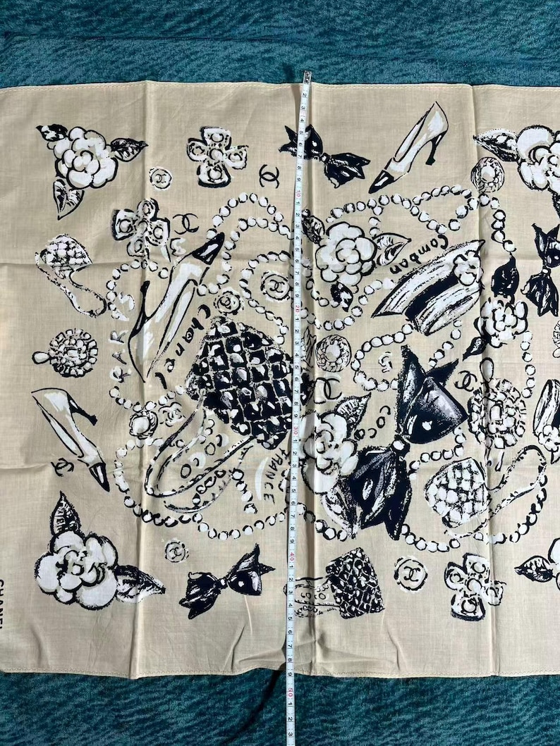 May include: Beige scarf with a repeating pattern of black and white illustrations. The design includes high heels, camellias, bows, and pearl necklaces. The word "Chanel" is printed vertically on the left side. The scarf measures approximately 40 inches in length.