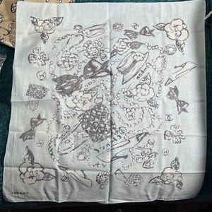 May include: A light blue square scarf with a repeating pattern of flowers, bows, and jewelry in black and gray. The word "FRANCE" is visible on the scarf. The brand name "CHANEL" is printed on the bottom.