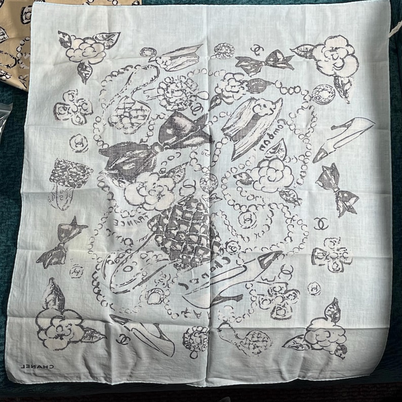 May include: A light blue square scarf with a repeating pattern of flowers, bows, and jewelry in black and gray. The word "FRANCE" is visible on the scarf. The brand name "CHANEL" is printed on the bottom.