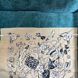 May include: Beige scarf with a black and white design featuring high heels, pearls, bows, and floral accents. The word "Chanel" is visible. The scarf is made of a soft fabric and is likely a fashion accessory.