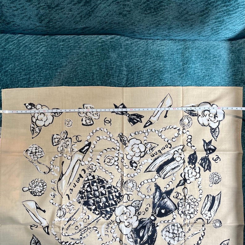 May include: Beige scarf with a black and white design featuring high heels, pearls, bows, and floral accents. The word "Chanel" is visible. The scarf is made of a soft fabric and is likely a fashion accessory.