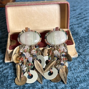 May include: A pair of vintage earrings with oval, striped, pale green and white stones. The earrings feature a beaded border, dangling beads, gold-toned leaf-shaped charms, and white circular charms. They are displayed in a small, open, vintage box.