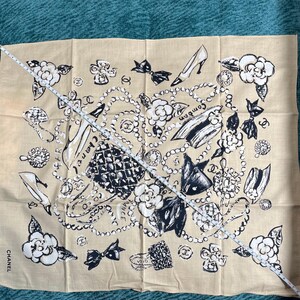 May include: Tan scarf with a black and white pattern featuring high heels, bows, camellias, and jewelry. The word "Chanel" is printed on the bottom left corner. The scarf is approximately 30 inches long.