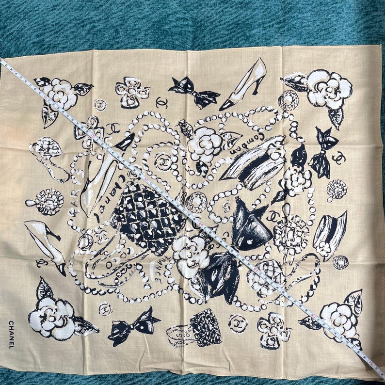 May include: Tan scarf with a black and white pattern featuring high heels, bows, camellias, and jewelry. The word "Chanel" is printed on the bottom left corner. The scarf is approximately 30 inches long.