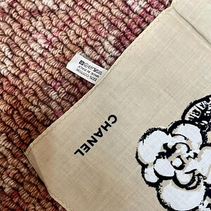 May include: Beige cotton handkerchief with a black and white floral design and the brand name "CHANEL" printed in black. The handkerchief has a tag that reads "100% Cotton Made in Italy".