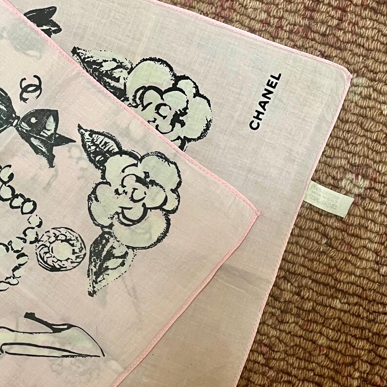 May include: Two pink handkerchiefs with black and white illustrations. One features a bow, jewelry, and a shoe, while the other displays a flower and the word "CHANEL". Both have pink borders.