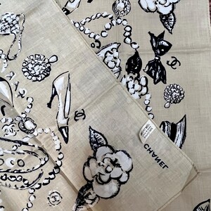 May include: Beige cotton handkerchief with black and white illustrations of high heels, jewelry, camellias, and bows. The word "CHANEL" is printed on the handkerchief. The handkerchief is folded in half.