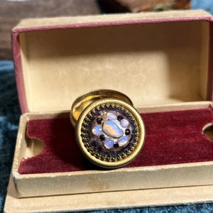 Peut inclure: Une bague vintage dorée avec un motif circulaire orné. La bague présente un groupe central de pierres opalescentes entouré d'une bordure violet foncé et d'un bord doré décoratif. Elle repose dans une petite boîte vintage ouverte avec un intérieur en velours rouge.