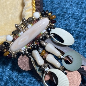 May include: A detailed, ornate brooch with a central oval, cream-colored stone. The brooch is surrounded by a variety of beads in black, gold, brown, and white. Additional decorative elements include cream-colored teardrop shapes and circular accents.