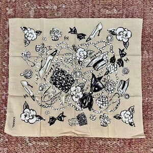 May include: Beige square scarf with a black and white design featuring high heels, camellias, bows, and jewelry. The word "CHANEL" is printed on the bottom left corner. The scarf is made of a lightweight fabric.