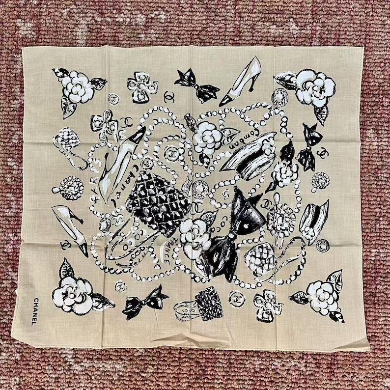 May include: Beige square scarf with a black and white design featuring high heels, camellias, bows, and jewelry. The word "CHANEL" is printed on the bottom left corner. The scarf is made of a lightweight fabric.