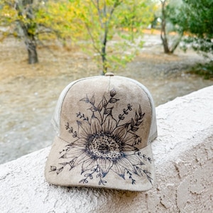 Könnte beinhalten: Beige Trucker-Cap mit detailliertem schwarzem Sonnenblumen-Design auf der Vorderseite. Die Kappe hat eine Mesh-Rückseite und einen gebogenen Schirm. Die Sonnenblume ist von Blättern und Stängeln umgeben.