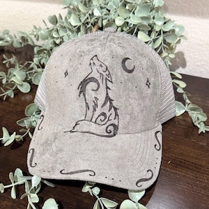 Puede incluir: Gorra de camionero gris con un diseño de lobo aullando. La gorra presenta una parte trasera de malla, una visera curva y una ilustración detallada de un lobo, una luna creciente y estrellas. El diseño del lobo es negro y la gorra tiene remolinos decorativos.