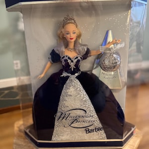 May include: A Barbie Millennium Princess doll in a clear display case. The doll wears a dark blue gown with silver accents, a tiara, and a silver necklace. The packaging reads "Special Millennium Edition" and "Millennium Princess Barbie."