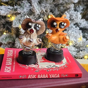 May include: Two whimsical owl figurines with large eyes, wearing hats, and attached to springs. One is brown and white, the other orange. They sit on a red book titled "ASK BABA YAGA" and a dark purple book.