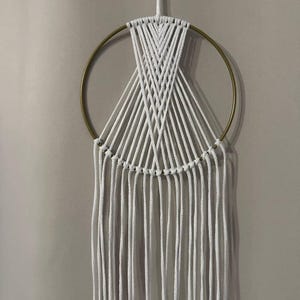 Könnte beinhalten: Handgefertigter Makramee-Wandbehang mit einem goldenen Metallring und weißer Baumwollschnur. Das Design umfasst ein gewebtes Muster und lange, vertikale Fransen. Diese Dekoration im Bohème-Stil verleiht jedem Raum Textur und Kunst.