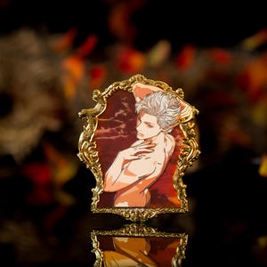 Puede incluir: Un pin de metal dorado que presenta una ilustración enmarcada de una persona con cabello blanco y torso desnudo. El marco tiene detalles ornamentados y un fondo rojo y marrón. El pin está sobre un fondo borroso con colores otoñales.