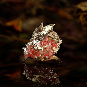Puede incluir: Broche metálico dorado con un diseño detallado de una figura con casco y cabello rojo. El broche tiene un acabado brillante y se encuentra sobre un fondo oscuro y borroso con hojas de otoño.