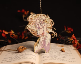 May include: A gold-colored pendant featuring an anime-style illustration of a figure in a flowing gown, suspended in front of an open book. The figure is surrounded by ornate, swirling details. The background is dark with autumnal foliage.