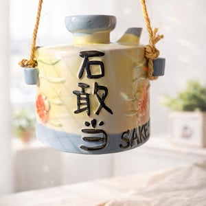 以下が含まれることがあります： 黄色と青色の陶器製酒ディスペンサー。日本語の文字と花の模様が施されています。ロープで吊り下げられており、注ぎ口があります。「SAKE」の文字が側面に印刷されています。