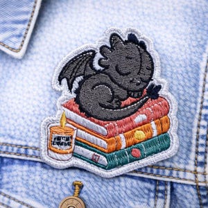 May include: Embroidered patch featuring a sleeping black dragon atop a stack of colorful books. A small candle with text is next to the books. The patch is sewn onto a light blue denim jacket.
