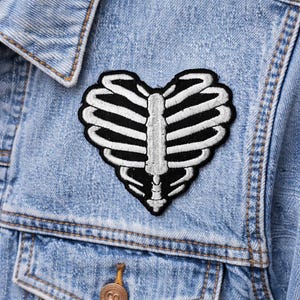 May include: A black and white heart-shaped patch featuring a rib cage design. The patch is embroidered with black and white thread and is affixed to a light blue denim jacket. The patch is a unique accessory.