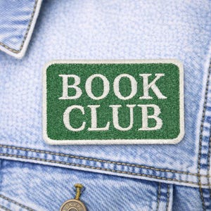 May include: A green rectangular patch with the words "BOOK CLUB" embroidered in white. The patch is sewn onto a light blue denim jacket. The jacket has visible stitching and a button.