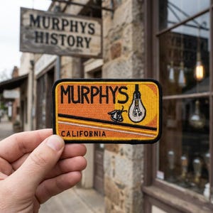 May include: Rectangular patch with the word "MURPHYS" in black letters on an orange background. A lightbulb and a moth are also featured. The word "CALIFORNIA" is at the bottom. The patch is held in front of a building with a sign that says "MURPHYS HISTORY".