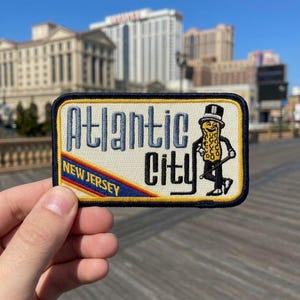 May include: Embroidered patch featuring the text "Atlantic City, New Jersey" with a cartoon peanut character in a top hat and cane. The patch has a cream background with blue and yellow accents and a black border.