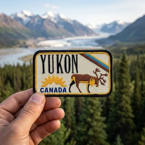 May include: A rectangular patch with the word "YUKON" in black letters, a yellow sunburst, and a brown caribou. The word "CANADA" is below the sunburst. The patch has a black border and a cream background.