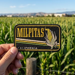 May include: A rectangular patch with the word "MILPITAS" in yellow lettering above a corn graphic. Below the corn is a horizontal stripe design and the word "CALIFORNIA". The patch is black with yellow and green accents.