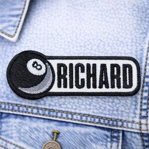 May include: Embroidered patch featuring a black and white 8-ball design and the name "RICHARD" in bold, black letters. The patch is rectangular with rounded corners and is affixed to a light blue denim jacket.