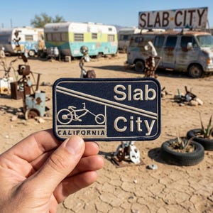 May include: A navy blue patch with the words "Slab City" in white, along with a bicycle graphic and the word "California". The patch is held in front of a desert landscape with vintage RVs and a sign that reads "Slab City".