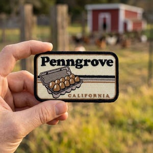 May include: A rectangular patch with the word "Penngrove" in black, above an illustration of a carton of eggs. The patch also includes the word "CALIFORNIA" in black. The patch is beige with a black border and brown stripes.