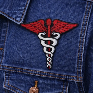 May include: A denim jacket with a red and white medical symbol patch. The patch features a staff with wings and a serpent, a common symbol in healthcare. The jacket has a classic design with button closures and pockets.
