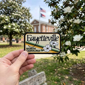 May include: A rectangular patch with the word "Fayetteville" in a cursive font, with "North Carolina" below. The patch features a green and yellow stripe design and white flower illustrations. The patch is held in front of a blurred outdoor background.