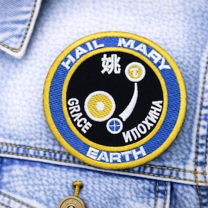 May include: Embroidered patch with the words "HAIL MARY GRACE EARTH" in a circular design. The patch features a black center with yellow and blue borders, and includes Chinese and Cyrillic text. It is attached to a denim jacket.