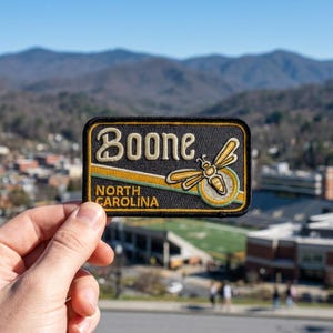 May include: A rectangular embroidered patch with a black background featuring the text Boone and North Carolina in cream lettering, accented by a stylized yellow and cream bee graphic and a diagonal yellow and cream stripe across the center.