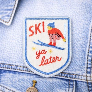 May include: Embroidered patch with a skier design and the words "SKI ya later." The patch features a skier in red and blue on a white background with a blue border. The patch is sewn onto a light blue denim jacket.