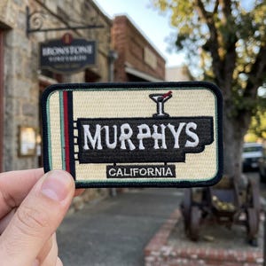 May include: Embroidered patch featuring the word "Murphys" in white lettering on a black background, with a cocktail glass icon. The patch also includes the text "California". The patch has a cream-colored background with green and red stripes.
