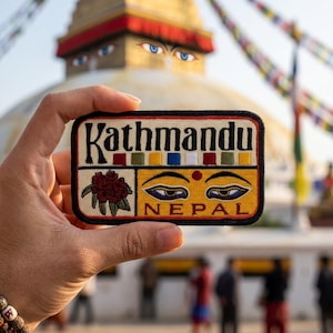 May include: Rectangular patch with the word "Kathmandu" in black letters. The patch has a red, yellow, blue, and white design. Below the word "Kathmandu" is a design of eyes and the word "NEPAL".