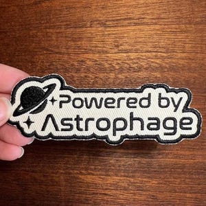 May include: A rectangular patch with the text "Powered by Astrophage" in black, outlined in black, on a cream-colored background. The patch features a black planet graphic with a ring and two small star graphics.