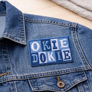 May include: A blue denim jacket with a rectangular patch that reads "OKIE DOKIE" in white and blue lettering. The patch is sewn onto the jacket's front, near the chest pocket. The jacket has a button closure and a classic collar.