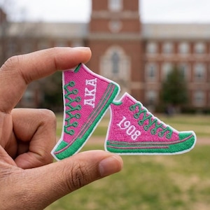 May include: Two pink and green embroidered sneaker patches. One patch has the letters "AKA" in green, and the other has the number "1908" in white. The patches are held up in front of a blurred background of a building.