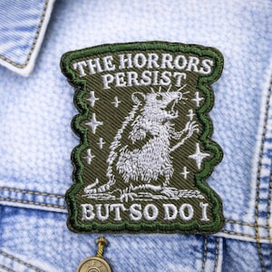 May include: Embroidered patch with the text "THE HORRORS PERSIST BUT SO DO I" and a detailed illustration of a rat. The patch is olive green with white and black accents, and is sewn onto a light blue denim jacket.