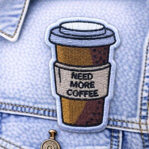 May include: Embroidered patch featuring a coffee cup design with the text "NEED MORE COFFEE." The patch has a blue, brown, and tan color scheme and is attached to a denim jacket.